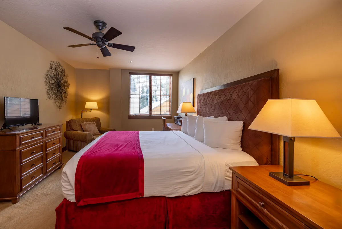 This image depicts a king-sized bed adorned with a red throw, a wooden dresser, a comfortable chair, and a window letting in natural light. The overall feeling is restful and inviting, suggesting a serene retreat for relaxation after a day of mountain adventures.