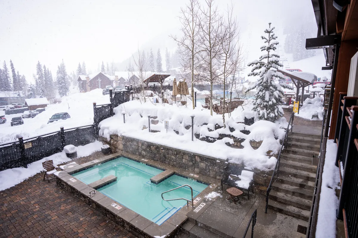 An inviting winter scene of an outdoor hot tub surrounded by snow-covered trees and landscape. The steps lead down to the tub, which steams invitingly against the crisp mountain air, offering a tranquil space for guests to relax after a day on the slopes.