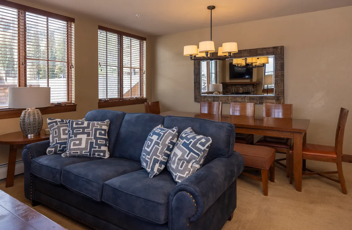 A welcoming dining and living area with a stylish blue sofa adorned with decorative pillows, a dining table ready for intimate meals or games, a modern light fixture, and a wall mirror that reflects the light and space. The scene exudes comfort and the joy of gathering after a day exploring the village.