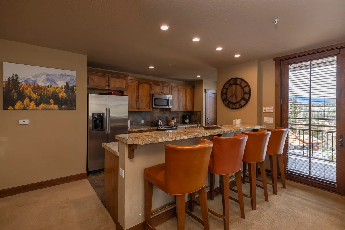The image focuses on the kitchen area, featuring a granite countertop with vibrant orange bar stools, modern appliances, and a large window that offers a scenic mountain view. The kitchen feels both functional and inviting, perfect for preparing meals and enjoying morning coffee while soaking in the alpine scenery.