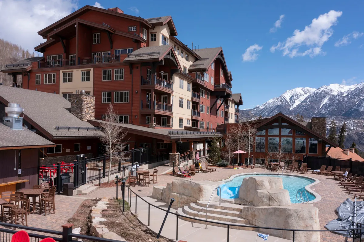 This image showcases the inviting outdoor pool area, with a clear view of the expansive mountains in the backdrop. The building facade, comfortable outdoor seating, and the serene pool all highlight the luxurious and family-friendly amenities available for guests. The setting transitions beautifully from summer sunbathing to winter hot tub relaxation, reinforcing the year-round appeal of the property.