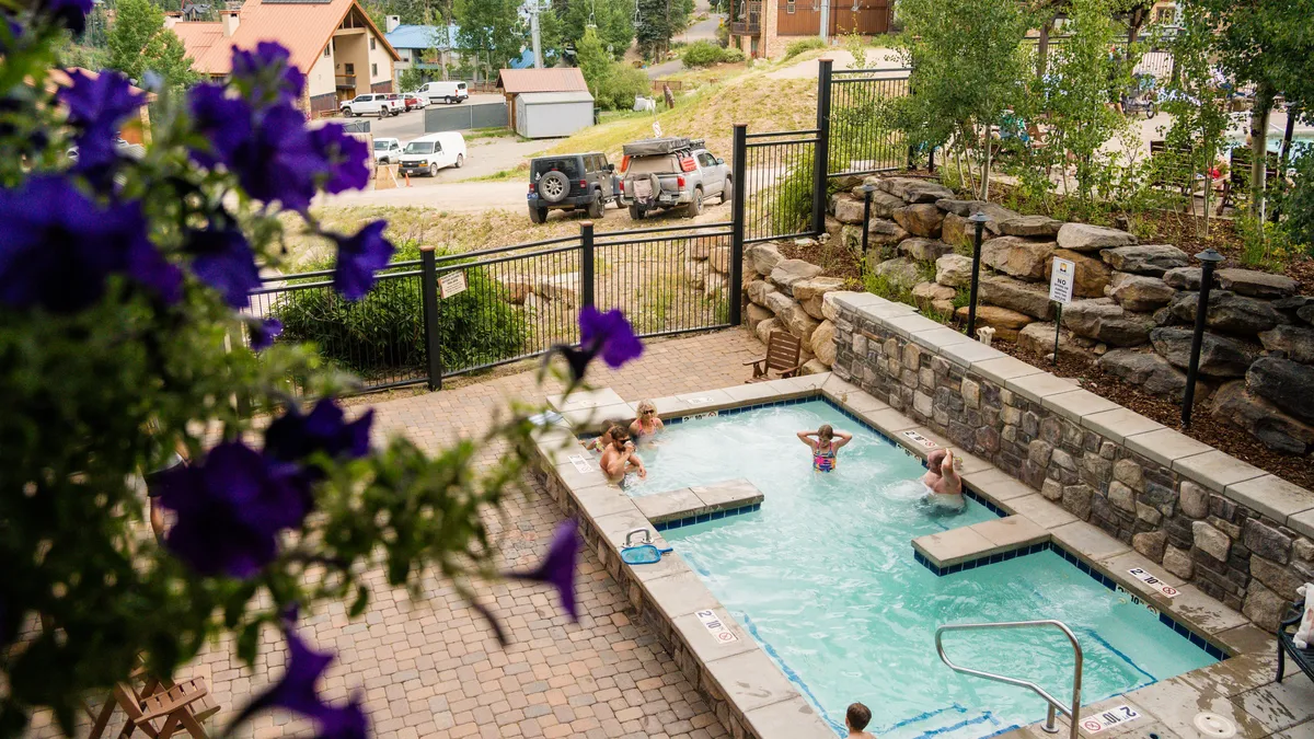 An inviting outdoor area featuring a bubbling hot tub and adjacent swimming pool, set amidst lush greenery and flowering plants. Guests are depicted relaxing in the hot tub and enjoying the serene outdoor space, suggesting a leisurely, social vibe.