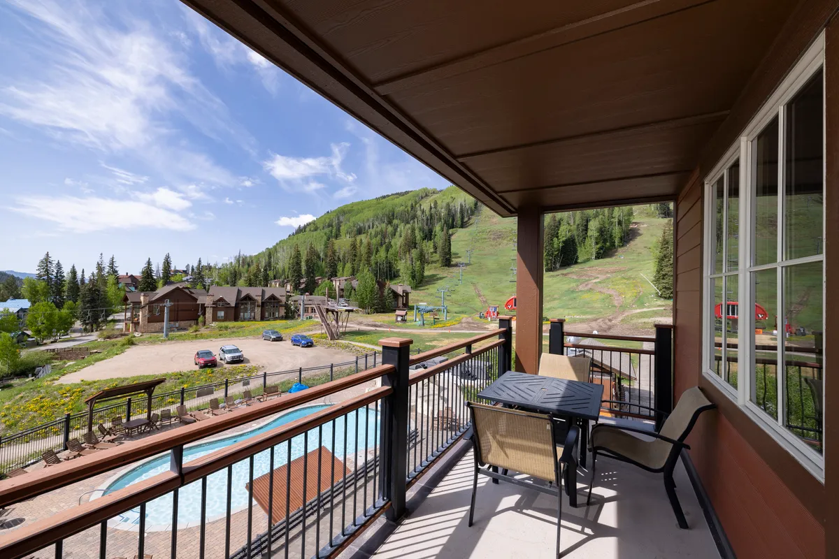 The image showcases the exterior of a luxury condo at Purgatory Lodge, featuring a spacious balcony with comfortable seating overlooking a stunning mountain vista. Guests can be seen relaxing on the balcony and socializing, while a sparkling swimming pool sits below, surrounded by lush evergreen trees. The tranquil atmosphere is heightened by the golden light of sunset casting a warm glow over the scene.