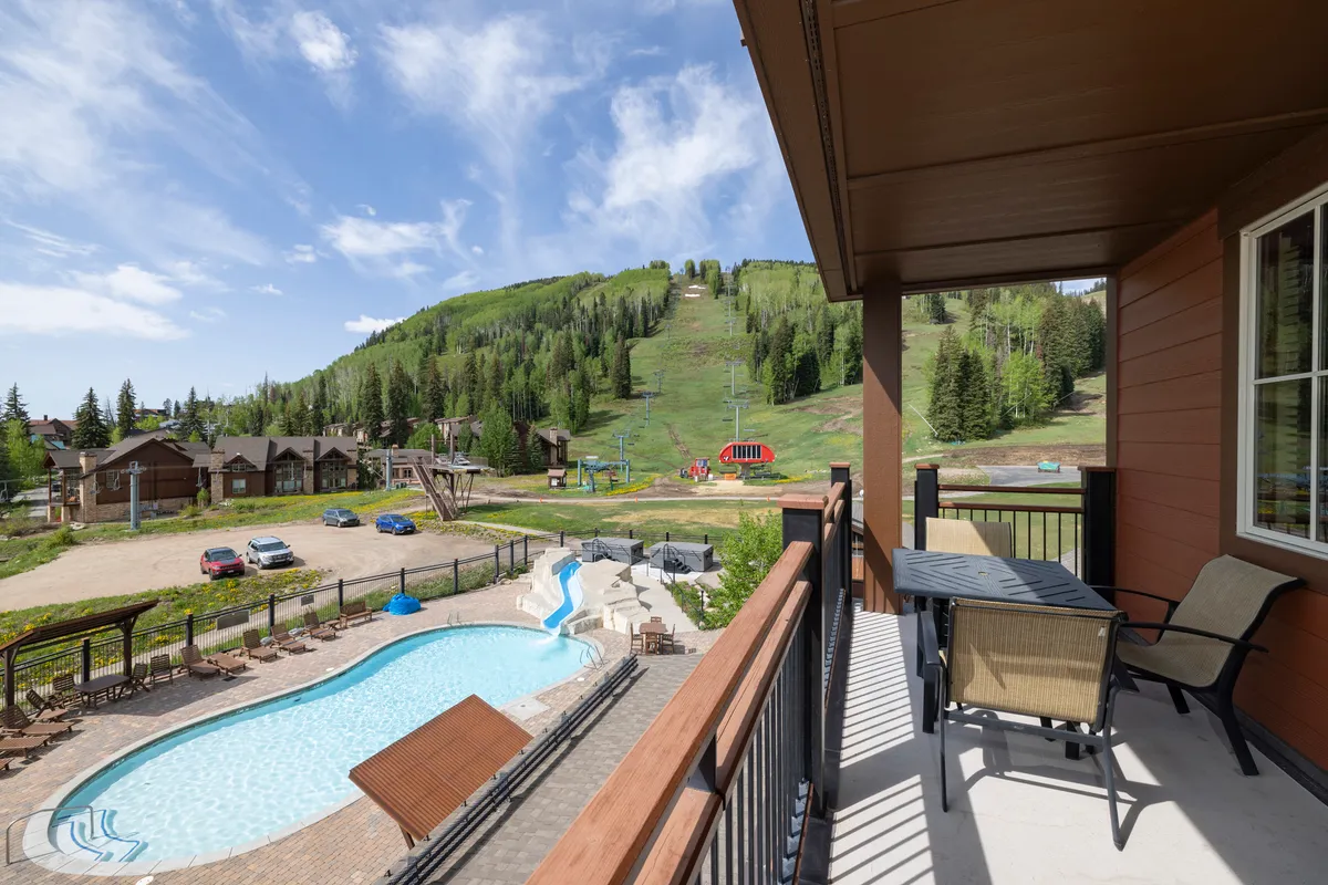 An inviting exterior scene featuring the lodge's heated swimming pool with steam rising, outdoor seating, and the ski slopes and lift in the background. The sun sets over the mountains, creating a beautiful contrast between relaxation and adventure. Guests are visible enjoying both poolside leisure and mountain activities.