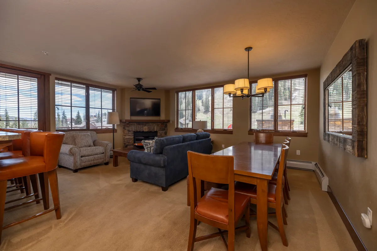 A cozy living and dining area is showcased, with a central fireplace, a comfortable sofa, a dining table perfect for family meals, and windows providing views outside. The setting is intimate and inviting, ideal for relaxation and lively conversation after a day on the slopes.