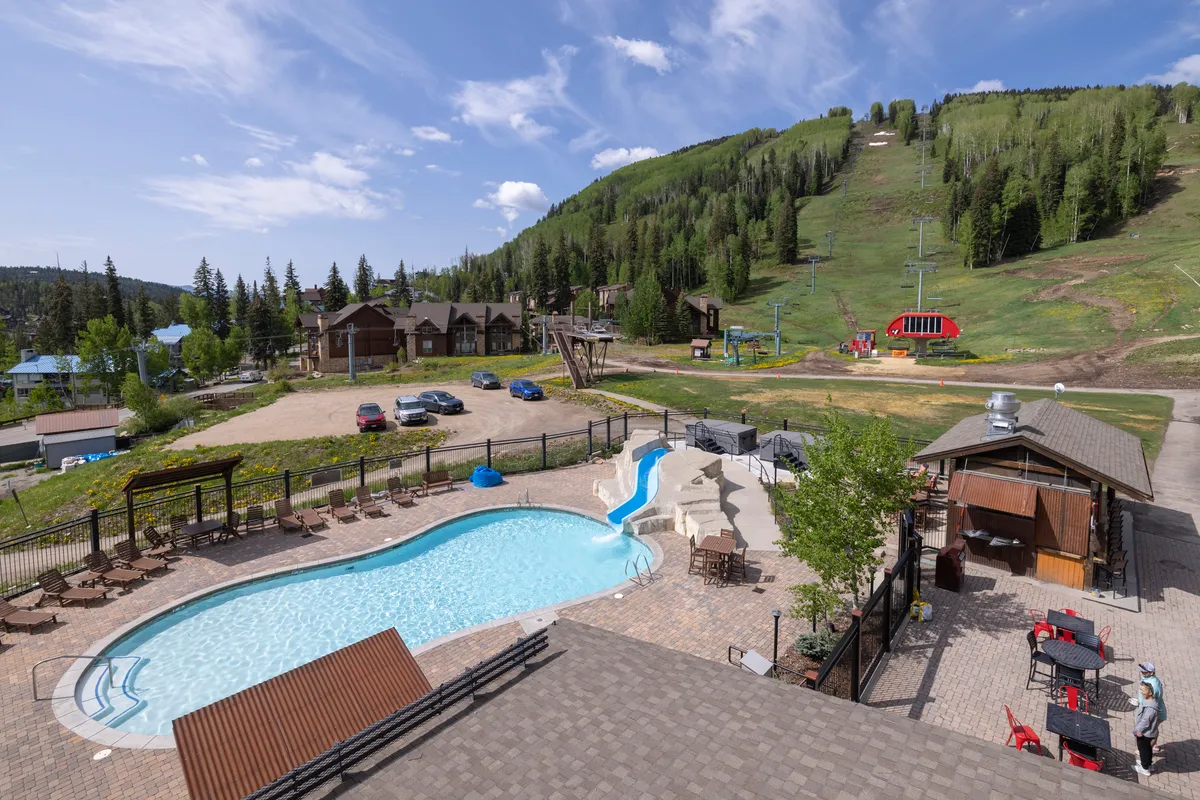 A vibrant outdoor pool scene featuring a large heated swimming pool with a prominent water slide curving into the water. Lounge chairs are arranged around the pool deck, and the area is surrounded by a stunning mountain backdrop. The atmosphere is lively yet relaxed, suggesting a resort-style communal space perfect for recreation and relaxation.