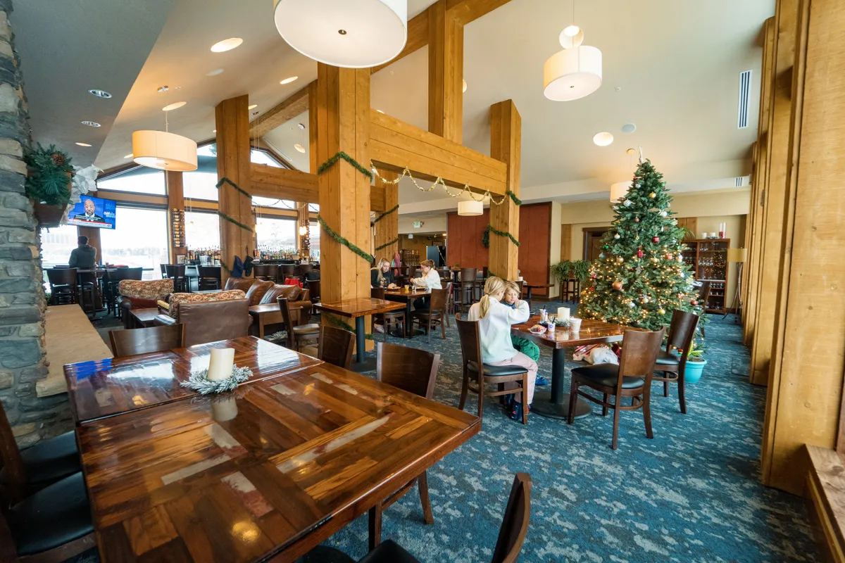 A festive restaurant or lounge setting featuring a decorated Christmas tree, wooden tables, a spacious bar area, and plenty of comfortable seating. Decorative lighting gives the space a warm, inviting feel, while the arrangement suggests a place for both casual dining and seasonal celebrations. The scene evokes a sense of gathering, socializing, and enjoying special moments together.