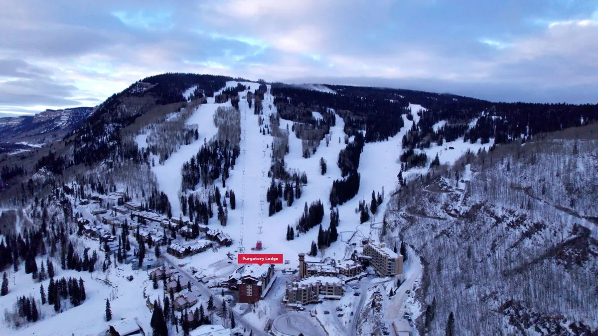 A sweeping exterior view of Purgatory Lodge nestled at the base of snow-covered ski slopes, surrounded by evergreen trees and with the majestic mountain landscape in the background. The lodge is seen welcoming guests as skiers traverse the runs right outside its doors, perfectly capturing the ski-in/ski-out concept.