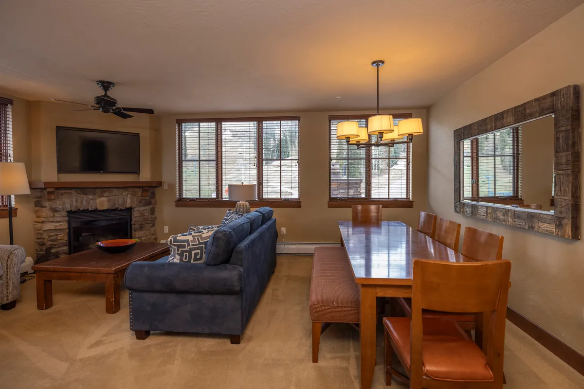 This image displays a spacious living area featuring a comfortable sofa, a dining table set for gatherings, a cozy fireplace, and large windows that bring in natural light and mountain views. The open layout seamlessly connects the living, dining, and kitchen spaces, creating a welcoming atmosphere for family and friends.