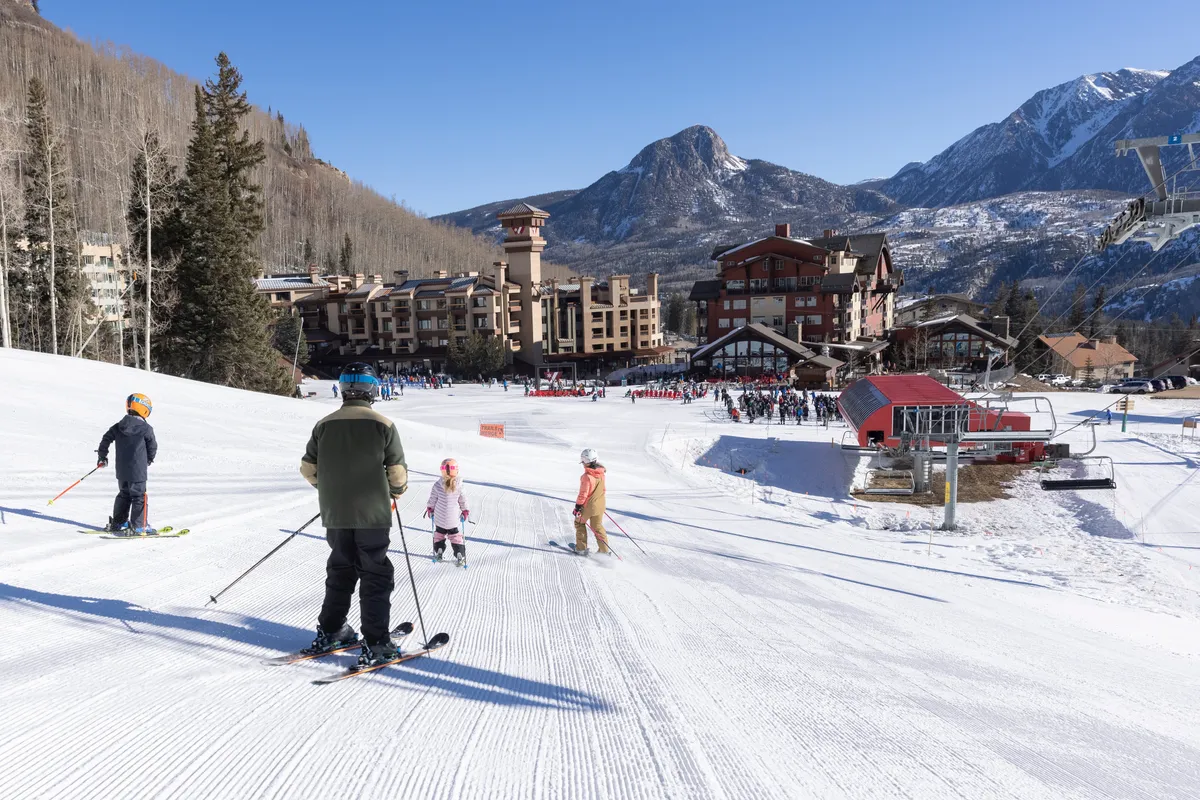 A lively scene outside the ski resort features skiers of various ages, the ski resort buildings, and the majestic mountain backdrop. The activity and visible resort entry tie directly into the ski-in/ski-out lifestyle and slope-side convenience.