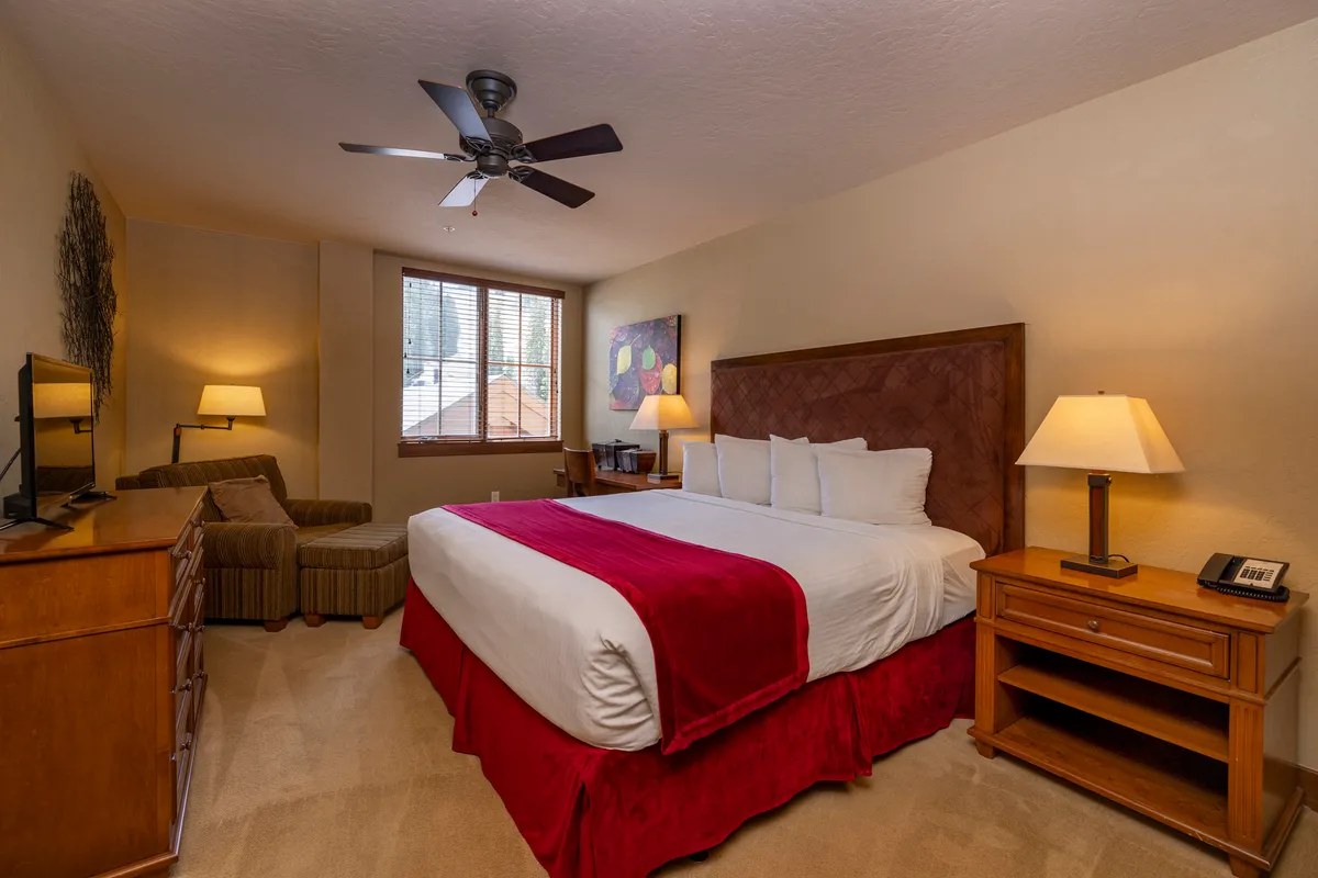 A cozy bedroom scene featuring a bed with red accents, wooden furniture, and a window that fills the room with natural light. The space feels comfortable and welcoming, ideal for unwinding.